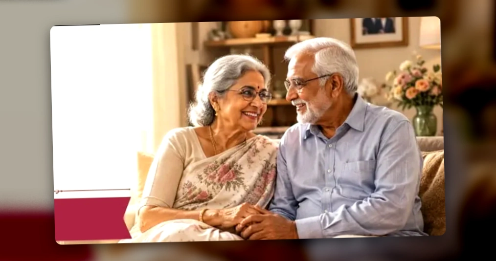 An elderly couple sits close together smiling on a sofa, with an anniversary cake and flowers on the table in front of them